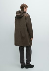 Brązowy wodoodporny płaszcz z kapturem, o luźnym kroju, z długimi rękawami i długością do połowy uda. Materiał o fakturze z przeszyciami w tonacji.