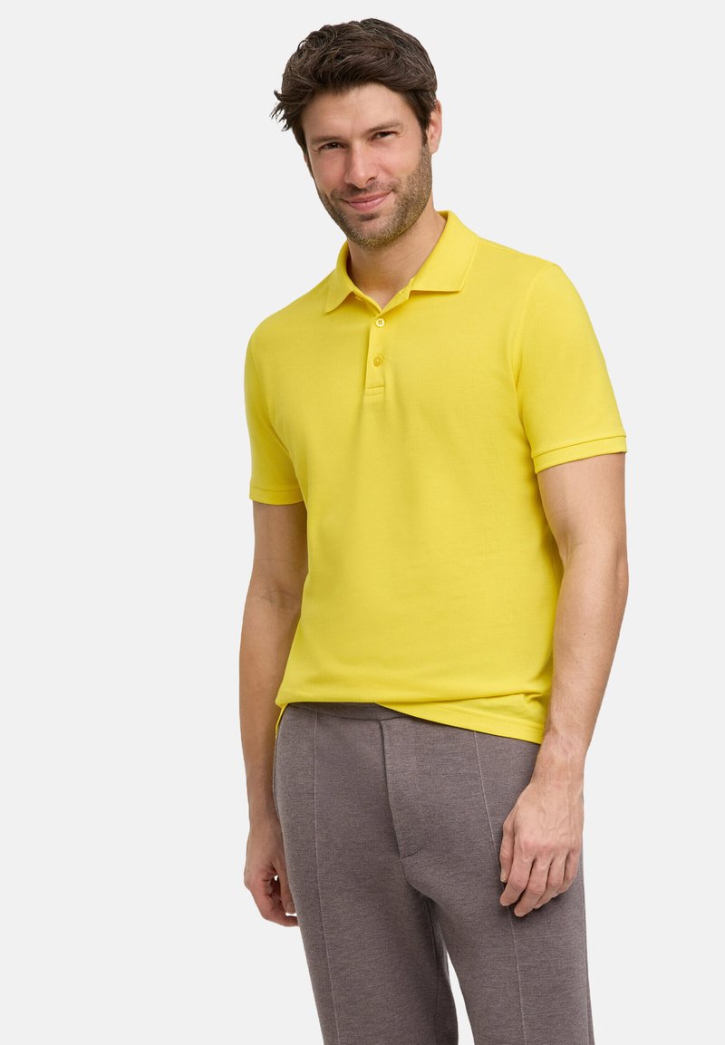 Żółta koszulka polo z gładkiego materiału, z klasycznym kołnierzykiem i zapięciem na dwa guziki. Połączona z szarymi, strukturalnymi spodniami.