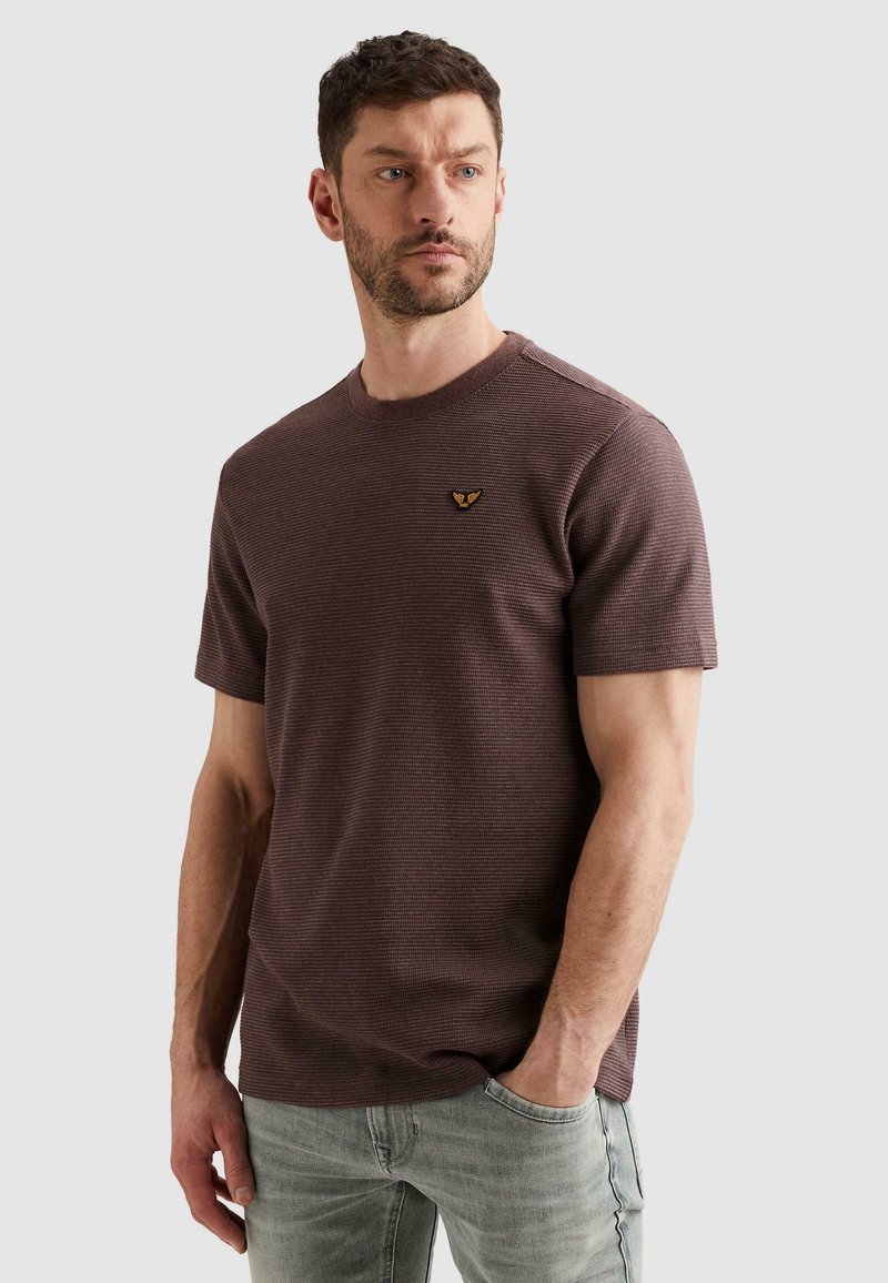 Homme aux cheveux courts et foncés avec une barbe, portant une chemise marron texturée à manches courtes et un jean gris clair, main dans la poche, regardant vers la gauche.