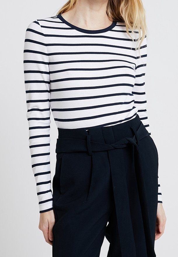 Femme portant une chemise blanche à manches longues avec des rayures horizontales noires et un pantalon noir taille haute avec une ceinture nouée.