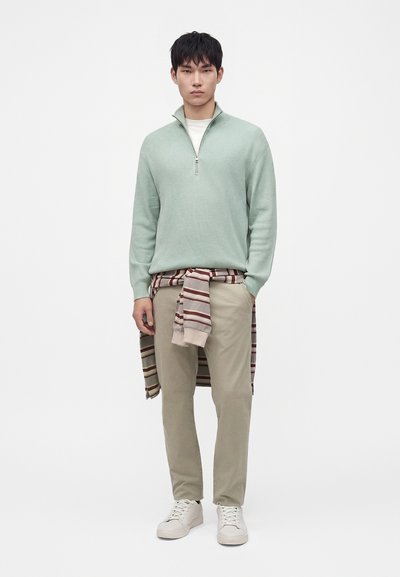 Jeune homme portant un pull vert clair à fermeture éclair au quart, un pantalon beige, des baskets blanches et un pull rayé beige et marron noué autour de sa taille.