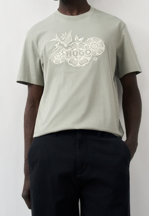 Homme portant un t-shirt gris clair avec un motif floral et d'oiseaux blanc entourant le texte "HUGO" et un pantalon noir, les mains dans les poches.