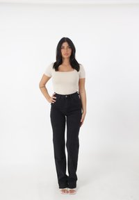 Top ajustado beige con escote cuadrado, combinado con jeans negros de cintura alta y sandalias de tacón transparente. Fondo blanco.