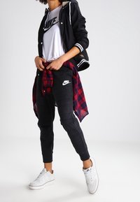 Chaqueta deportiva negra con rayas blancas, camiseta blanca de Nike, pantalones jogger grises y camisa a cuadros roja y negra atada a la cintura. Zapatillas blancas completan el conjunto.