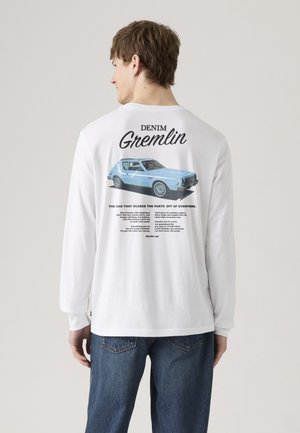 Moški v beli majici z dolgimi rokavi s tiskom starinskega modrega avtomobila in besedilom "Denim Gremlin" na hrbtu, v kombinaciji z modrimi jeans hlačami.