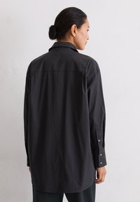 Vrouw met zwart haar in een bun, gekleed in een losse zwarte blouse met knopen, staat zijwaarts tegen een eenvoudige witte muur.