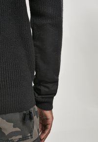 Suéter negro de punto con textura acanalada y tela negra acolchada en el codo. Lleva pantalones con patrón de camuflaje.
