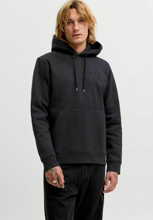 Mężczyzna z blond włosami do ramion ubrany w czarną bluzę Jack & Jones Premium i czarne dżinsy, stojący na tle jednolitego jasnego tła.