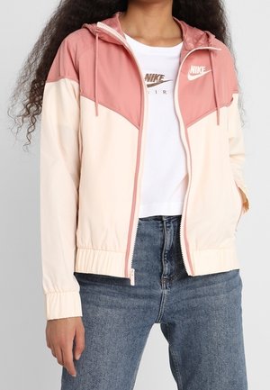 Person in einer rosa- und cremefarbenen Nike-Trainingsjacke über einem weißen Nike-Air-T-Shirt und blauen Jeans, mit einer Hand in der Jackentasche.