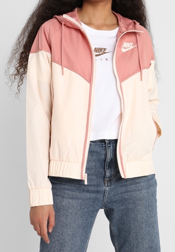 En person klädd i en rosa och krämfärgad Nike zip-up jacka över en vit Nike Air t-shirt och blå jeans, med en hand i jackfickan.