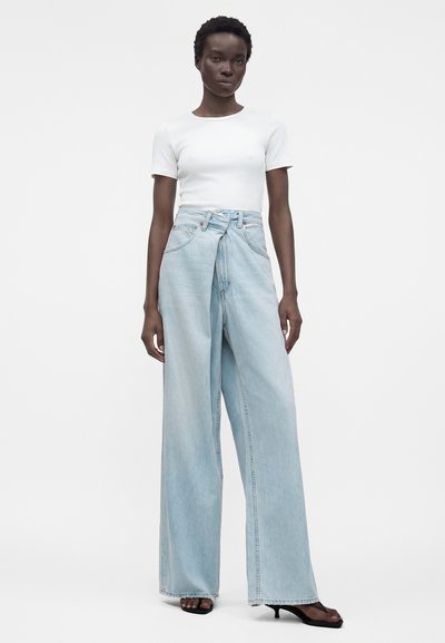 Rövid hajú nő, fehér, testhez simuló rövid ujjú felsőt, magas derekú, bőszárú világoskék farmert és fekete, sarkú szandált visel.