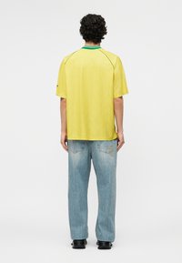 Hombre con cabello rizado de espaldas, vistiendo una camisa amarilla de manga corta con cuello verde y vaqueros azul claro con bolsillos traseros.