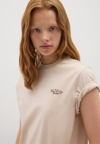 T-shirt en coton beige avec des manches courtes retroussées. Arbore l'inscription "SUNDAY Brunch" en cursive marron sur la zone du cœur gauche. Texture lisse.