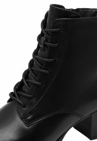 Botte noire en cuir au niveau de la cheville avec un bout arrondi, un laçage à l'avant et un talon empilé. Présente une texture lisse et des détails de surpiqûre subtils.