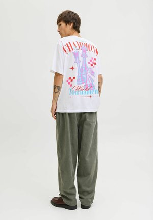Witte oversized t-shirt met kleurraf grafiek van schaakstukken en tekst op de achterkant. Combinatie met losse olijfgroene broek en donkere schoenen.