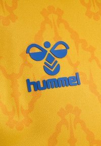 Μπλε λογότυπο Hummel με στυλιζαρισμένη μέλισσα πάνω από το μπλε κείμενο "hummel" σε κίτρινο υφασμάτινο φόντο με διακριτά πορτοκαλί μοτίβα.