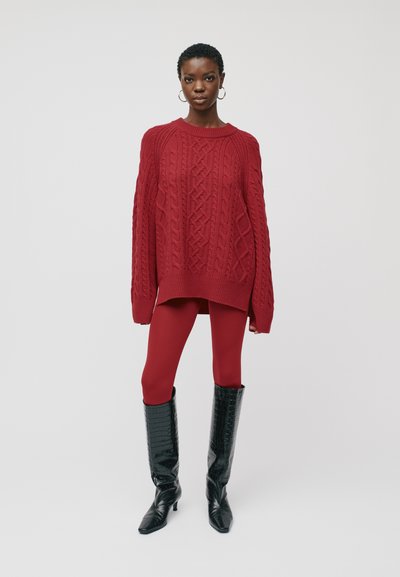 Pull en maille oversize rouge avec un design de câbles, associé à un legging rouge et des bottes noires brillantes montant jusqu'au genou avec des bouts pointus.