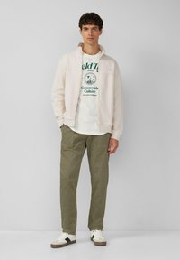 Hellbeige Zip-Up-Jacke über einem weißen T-Shirt mit grünem Aufdruck, kombiniert mit olivgrünen Cordhosen und weißen Sneakers mit schwarzen Akzenten.