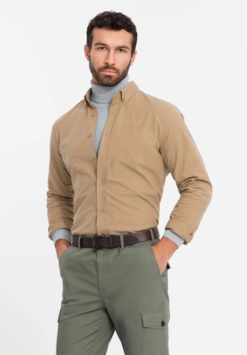 Beige button-up shirt met een borstzak, gecombineerd met een lichtgrijze turtleneck. Olijfgroene cargobroek met utilitaire zakken en een bruine riem.
