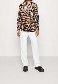 Button-up shirt in a tiger print pattern, featuring a silky texture in shades of tan and black, paired with white trousers and black shoes.