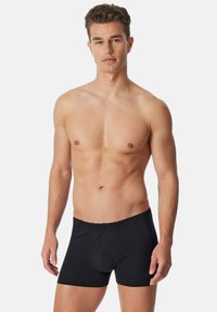 Sportig man med kort brunt hår iförd enkla svarta boxerkalsonger, stående mot en enkel ljusgrå bakgrund.