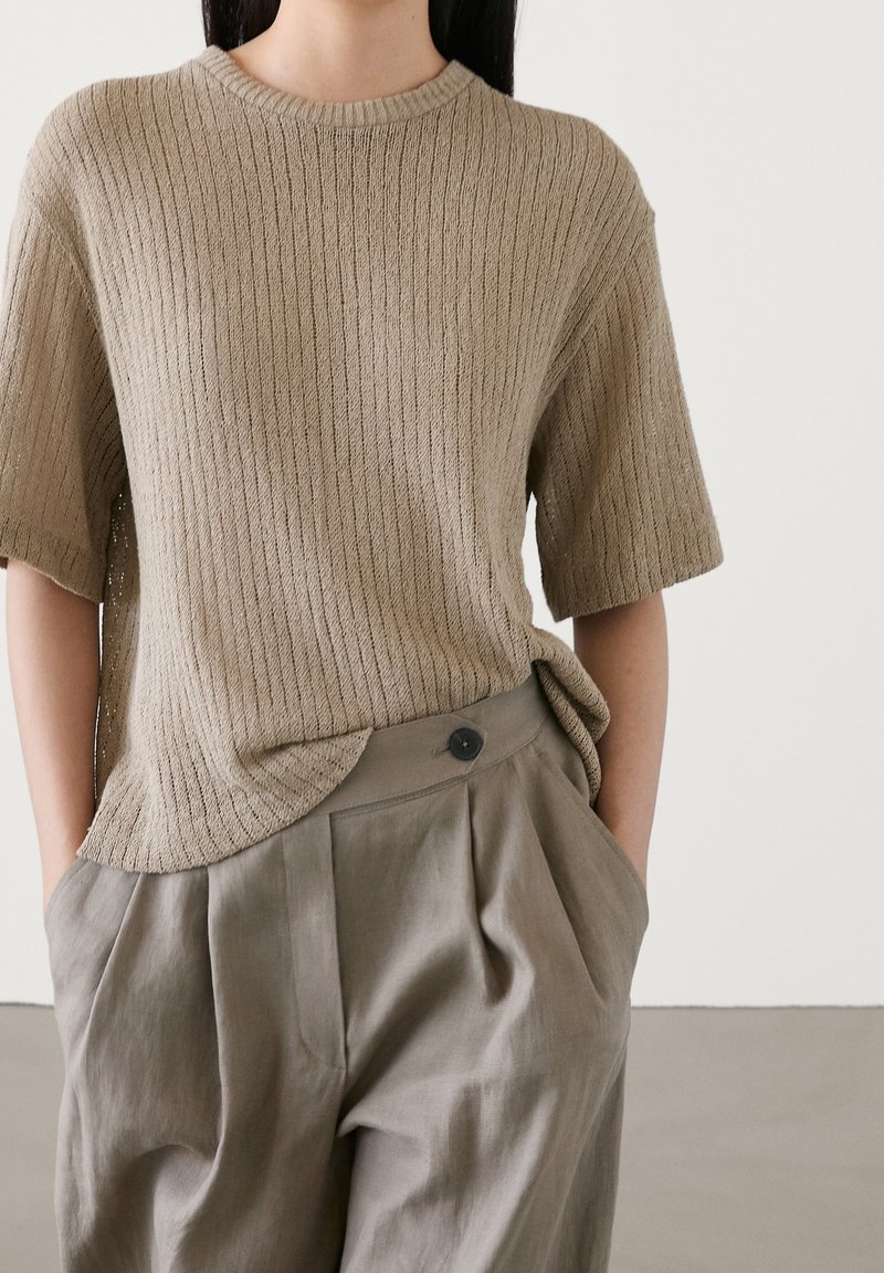 Beige ribbed knit top with short sleeves, round neckline, paired with loose olive-green pleated trousers featuring a single black button detail.