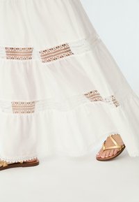 Robe maxi blanche avec des panneaux en dentelle, tissu fluide et design à niveaux. Des sandales avec des brides dorées complètent la tenue.