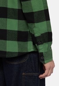Groene en zwarte geruite flanellen shirt met een geknoopte manchet, gemaakt van textuurstof. Donkerblauwe denim met zichtbare stikaccenten.