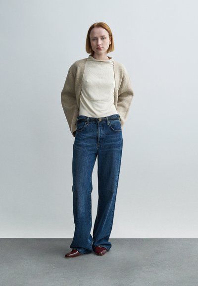 Μπεζ πλεκτό ζακέτα με υφή πάνω από μια κρεμ μπλούζα, συνδυασμένη με ψηλόμεσα μπλε τζιν και μπορντό παπούτσια. Απλές, καθαρές γραμμές σε όλο το σύνολο.