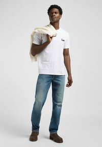 Camiseta blanca de algodón con el logo "Lee", jeans de mezclilla azul y botas marrones; una chaqueta de color claro colgada sobre el hombro. Fondo neutro.