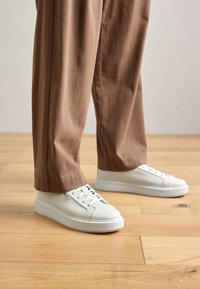 Persona che indossa sneakers bianche testurizzate a bassa altezza e pantaloni larghi marroni, in piedi su un pavimento in legno chiaro.