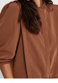 Blouse légère marron avec épaules froncées, col rond et coupe légèrement ample. Texture lisse avec des détails de panneaux latéraux.