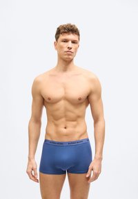 Jeune homme athlétique aux cheveux courts et bouclés, portant un boxer Björn Borg bleu, debout devant un fond clair uni.