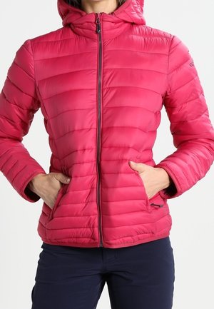 Femme portant une veste matelassée rose vif avec une fermeture éclair à l'avant et les mains dans les poches, associée à un pantalon foncé.