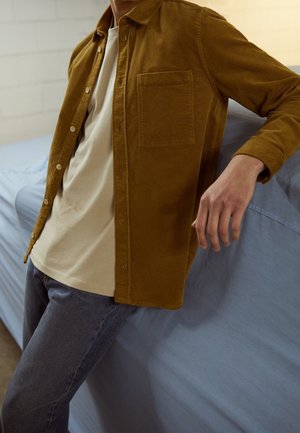 Homme appuyé sur un lit couvert de draps bleu clair, portant une chemise marron à boutons par-dessus un t-shirt beige et un jean foncé.