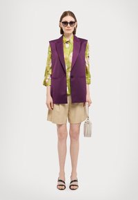 Lilla ermeløs blazer over en grønn blomstret skjorte, beige shorts og svarte sandaler. Holder en liten, lys håndveske.