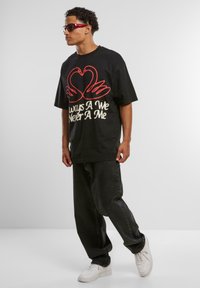 Man wearing black oversized T-shirt with red swan heart design and text, black baggy jeans, white sneakers, red sunglasses, standing on white floor.