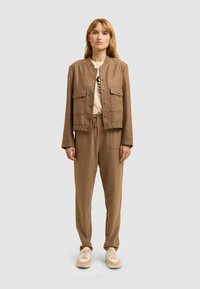 Femme se tenant de face, portant une veste marron avec des poches, un pantalon à cordon assorti, un t-shirt graphique blanc et des baskets beiges.
