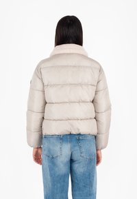 Giubbotto imbottito in beige chiaro con colletto in peluche, texture a righe orizzontali e polsini elasticizzati, abbinato a jeans blu sbiaditi.