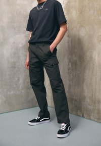 Person som bär svarta cargo-byxor, svart t-shirt med logo, svart och vit sneakers, och en silverkedja som står mot en betongvägg.