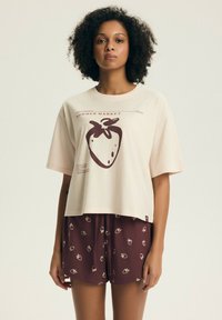 Kobieta o kręconych włosach, ubrana w beżowy t-shirt z dużym nadrukiem truskawki oraz pasujące brązowe szorty z małymi nadrukami truskawek.