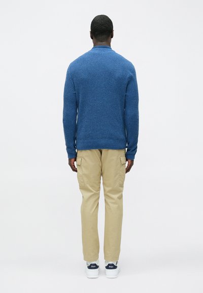 Mėlynas megztinis, smėlio spalvos karinių kelnių stiliaus kelnės, baltos sportinės avalynės. Dėmesys tekstūruotam mezgimo raštui, laisvam prigludimui ir praktiškoms kišenėms.