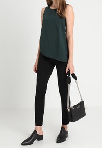 Ciemnozielony top bez rękawów, czarne dopasowane spodnie, czarne mule na słupku oraz mała czarna torebka z łańcuszkiem.