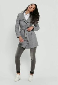 Cappotto in misto lana grigio con cintura a legare, chiusura con zip e tasche laterali, abbinato a pantaloni grigi e sneaker bianche. Texture liscia.