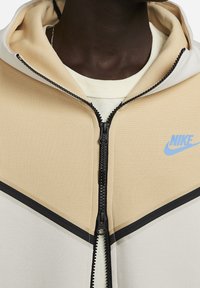 Zbliżenie na osobę w beżowej i białej bluzie z zamkiem Nike z niebieskim logo na piersi oraz w kremowej koszulce pod spodem.