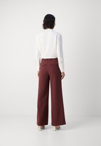 Valkoinen pitkähiha blouse, jossa on napitetut hihansuut, yhdistetty korkean vyötärön burgundinpunaisiin leveälahkeisiin housuihin, joissa on takataskut ja kultaiset yksityiskohdat.