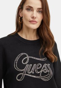 Felpa nera con collo rotondo, caratterizzata da un logo "Guess" in argento metallico decorato con strass. Texture morbida, design casual.