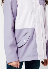 Fialová nepromokavá bunda s jemně fialovým panelem, s patentkami a šikmou přední kapsou. Hladká textura a volný střih.