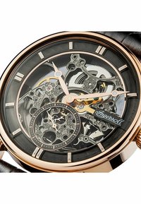 Skelettklocka med svart design, rosaguld accenter, läderrem, detaljerade kugghjul synliga och en liten sekundvisare.