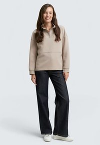 Pulover bej din fleece cu fermoar de un sfert, având un guler înalt și o cusătură orizontală. Asortat cu blugi largi de culoare albastru închis și pantofi albi.